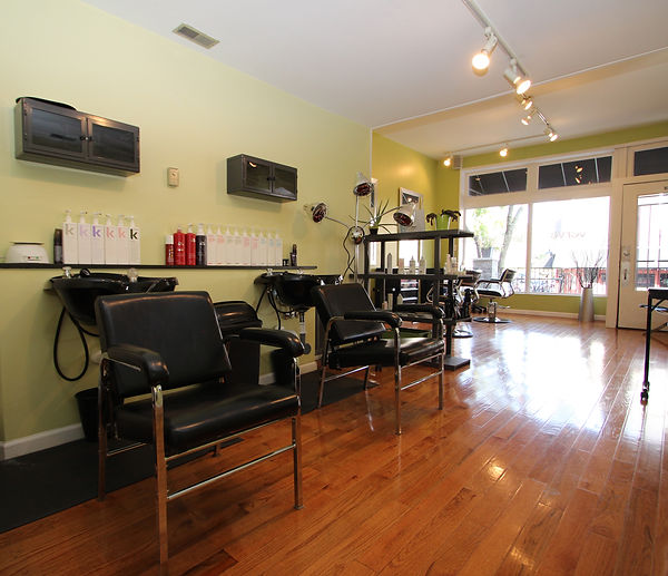 Shampoo bowls and black chairs along a green wall, with hardwood floors and front windows